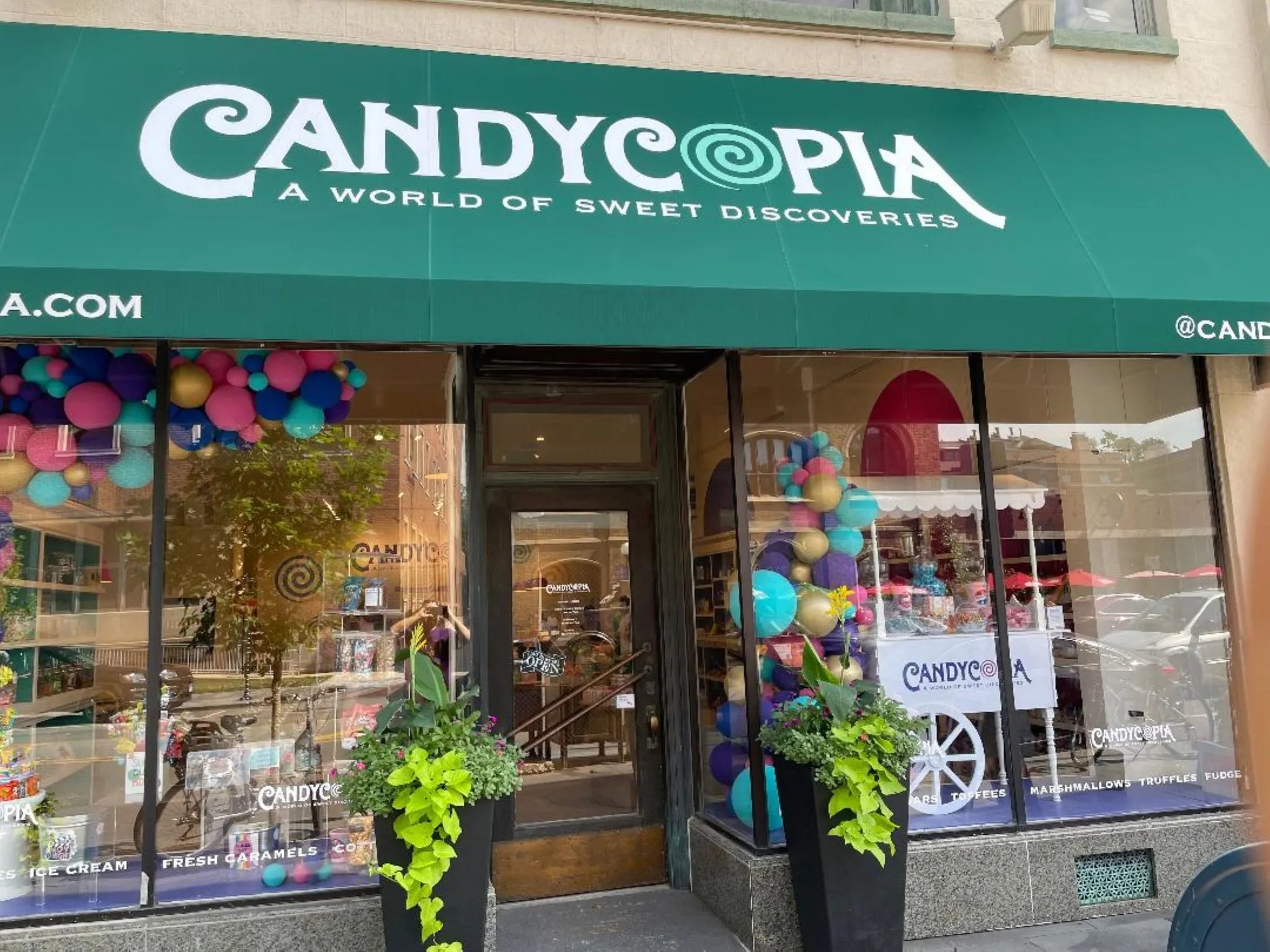 Candycopia storefront under a green awning reading CANDYCOPIA A WORLD OF SWEET DISCOVERIES, colorful balloon garlands and window displays