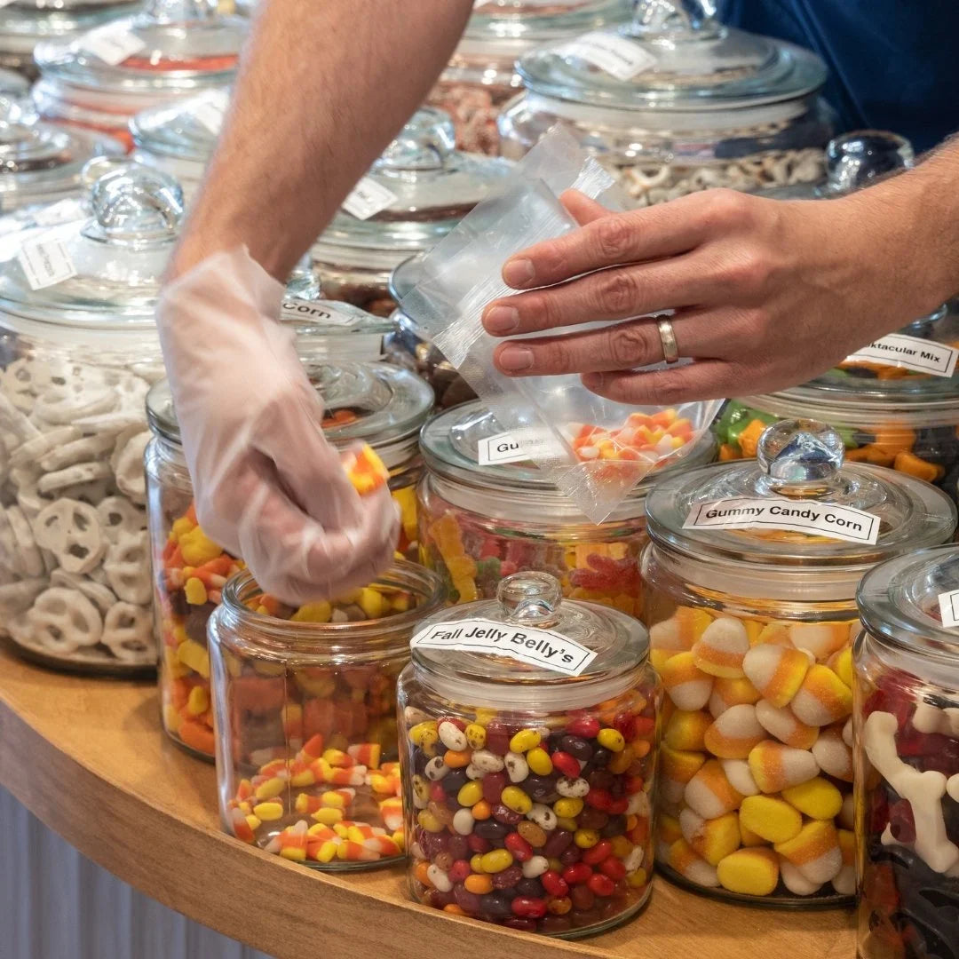 Hands scooping candy into a bag at a pick-and-mix counter with labeled glass jars of gummy candy corn and jelly beans