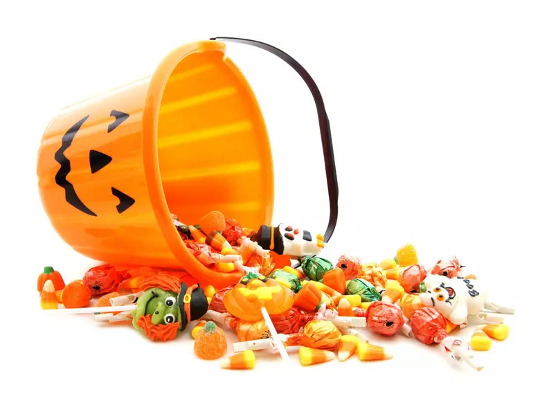 Jack-o’-lantern bucket tipped over spilling Halloween candy, including candy corn and wrapped lollipops, on a white background