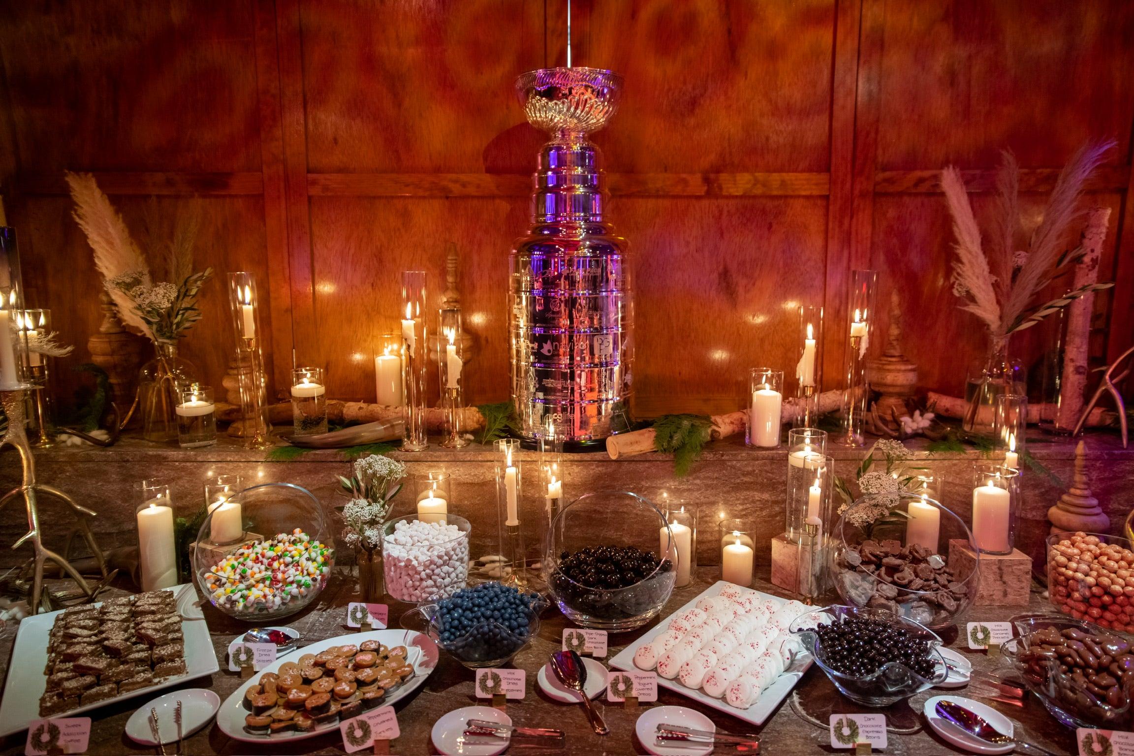 Pick-and-mix candy buffet display with glass bowls of assorted candies, cookies and meringues on a candlelit wooden table decorated with pampas grass