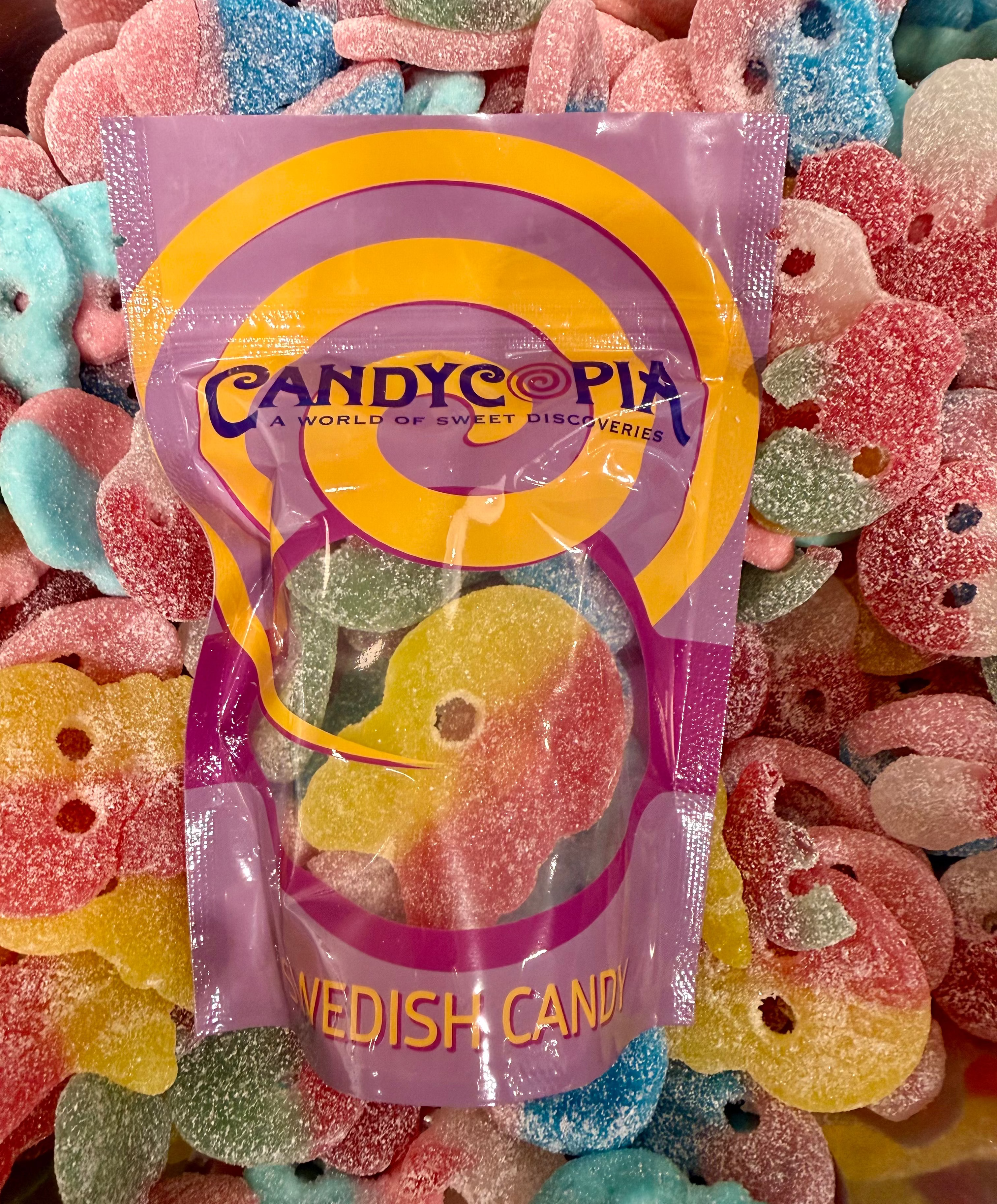 A bag of Candyco pia Swedish Candy sour gummy bears, shown in its purple and yellow packaging, surrounded by a pile of colorful, sugar-coated gummy skulls
