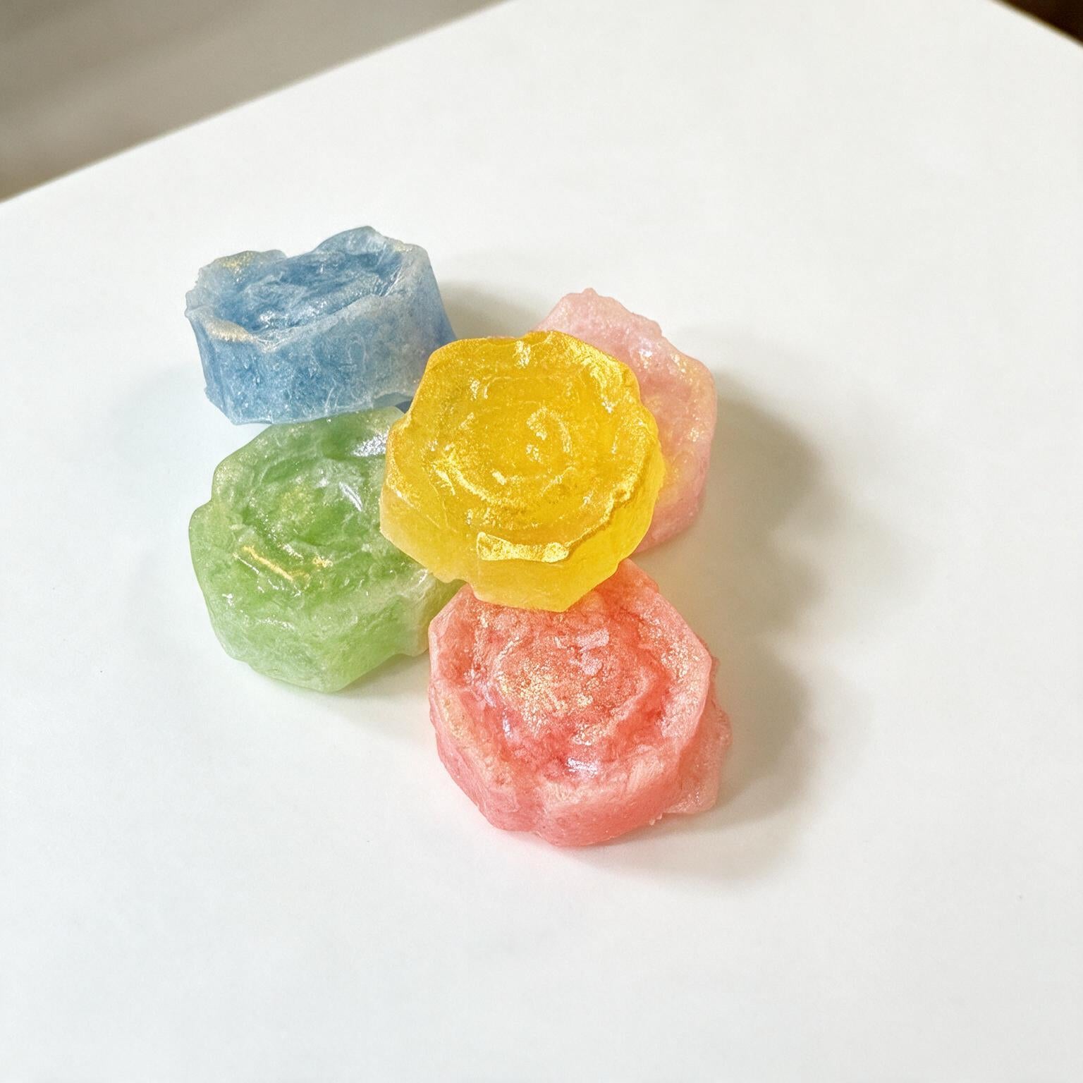 Four small translucent flower-shaped candies in blue, green, yellow, and pink arranged on a plain white surface
