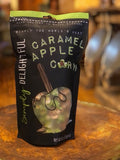 A black stand-up pouch of Simply Delightful Caramel Apple Corn, featuring a green and white apple graphic with a brown stick, set on a rustic wooden surface with a warm, blurred background