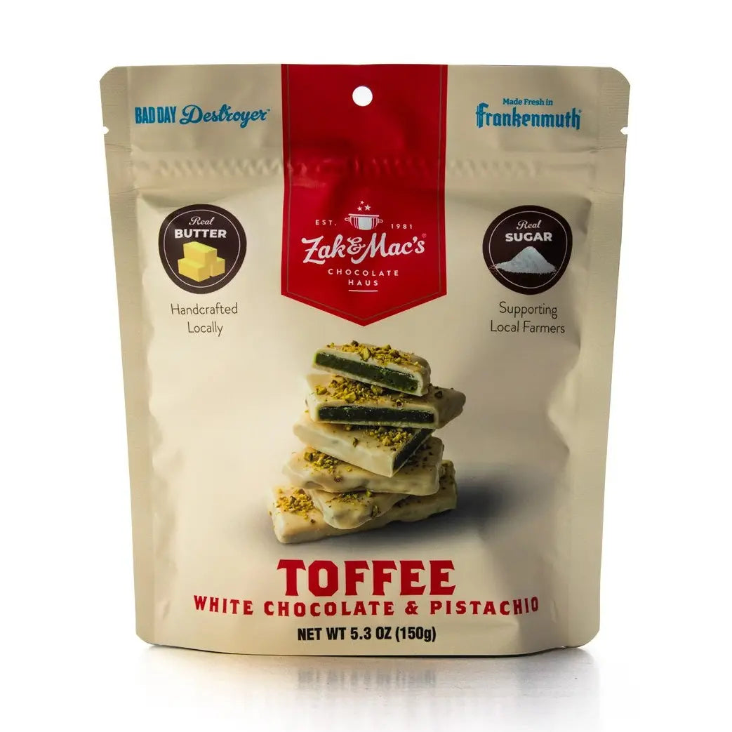 A stand-up pouch of Lake & Mac's Chocolate Works Toffee White Chocolate & Pistachio candies, featuring a beige background with a red central banner and an image of pistachio-topped white chocolate toffee pieces